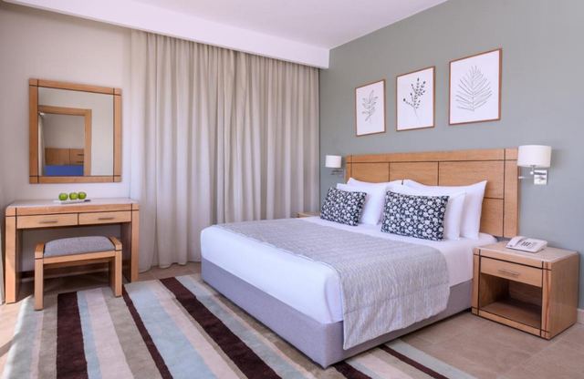 A hotel room: a grey-green wall with a light oak headboard, desk and mirror.